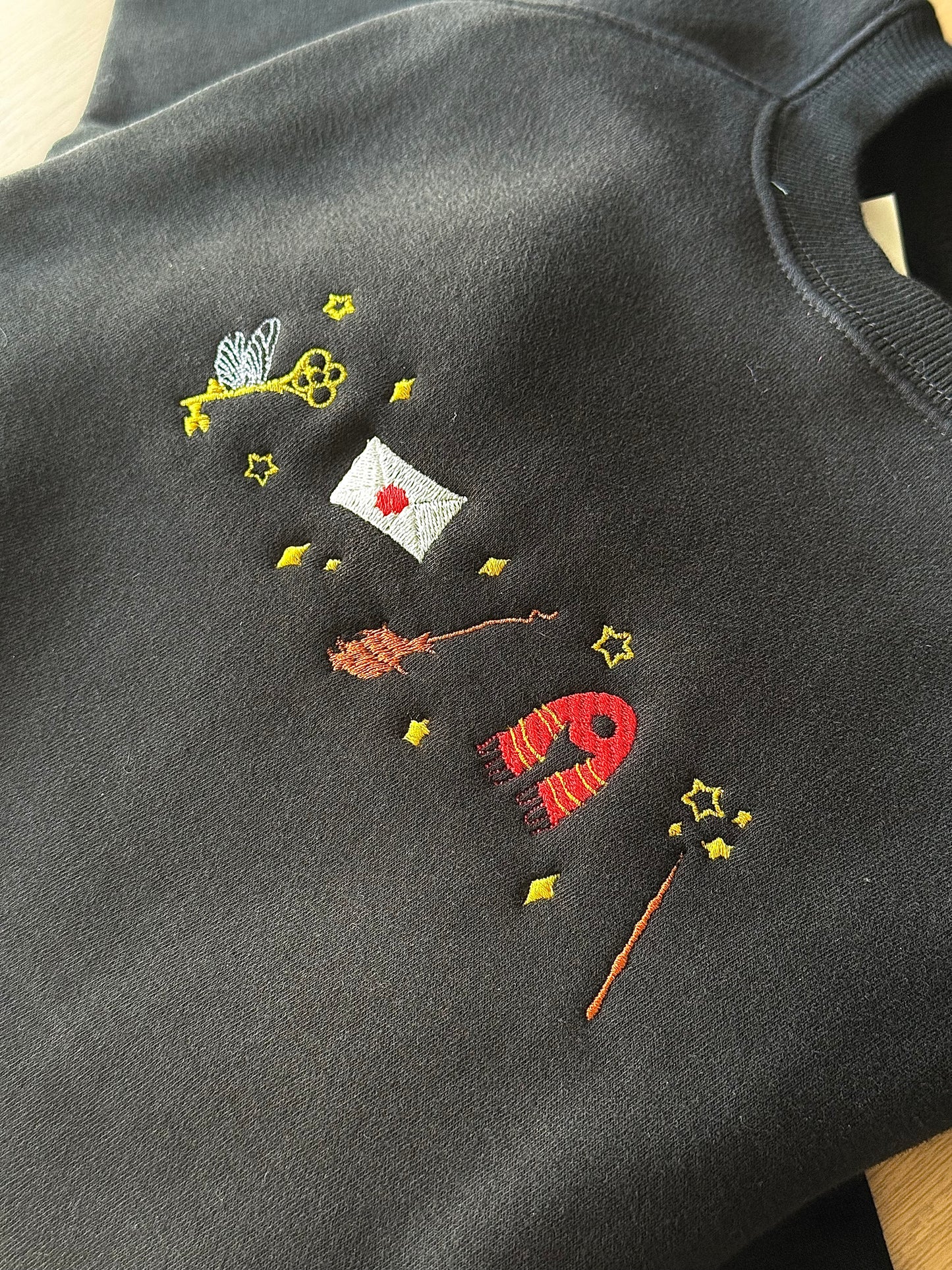 Embroidered design on a dark fabric with various elements including a key, envelope, and wand.
