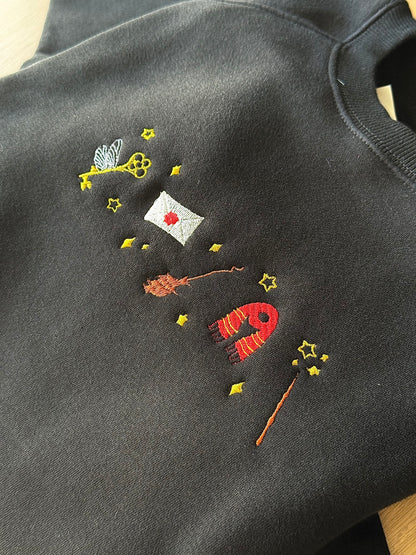 Embroidered design on a dark fabric with various elements including a key, envelope, and wand.